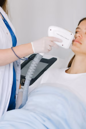 A medical professional in a white coat performing a facial treatment on a woman lying down, using a handheld device near her cheek.