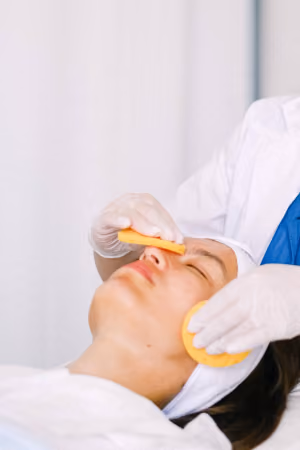 A person with eyes closed receiving a facial treatment while a professional wearing white gloves uses two orange sponges on the person's face in a clinical setting.