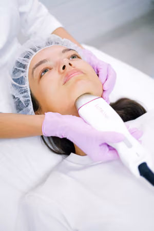 Woman lying down with a hair cover receiving a skin treatment using a handheld device on her neck by a person wearing purple gloves.