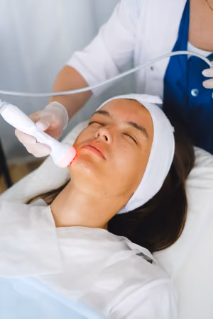 Woman lying on a treatment bed with eyes closed receiving a facial treatment using a handheld device emitting red light, by a person wearing white gloves and a white coat.