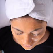 Top view of a person with medium skin tone lying down, wearing a white towel turban on their head and a black shirt, with a facial mask cream applied on their cheeks and forehead.