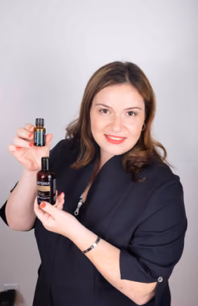 Smiling woman with shoulder-length brown hair and red lipstick holds up two dark-colored bottles of essential oils or wellness products in front of a plain light gray background.