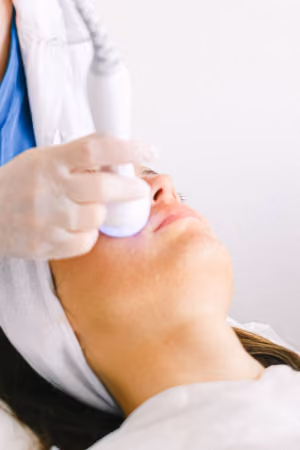 Woman receiving facial treatment with a handheld device emitting blue light, operated by a gloved professional wearing a white coat and head covering.