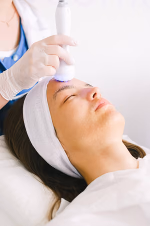 Woman lying down with eyes closed receiving a facial treatment using a handheld device by a gloved esthetician.