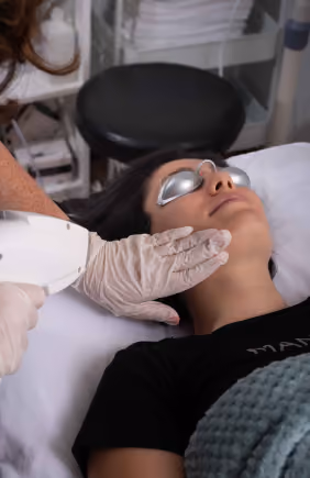 A woman lying on a treatment bed with protective goggles, receiving a facial laser treatment performed by a technician wearing gloves.