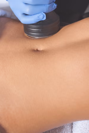 Close-up of a person’s bare abdomen with a gloved hand holding a round black device against the skin, suggesting a body treatment or therapy.