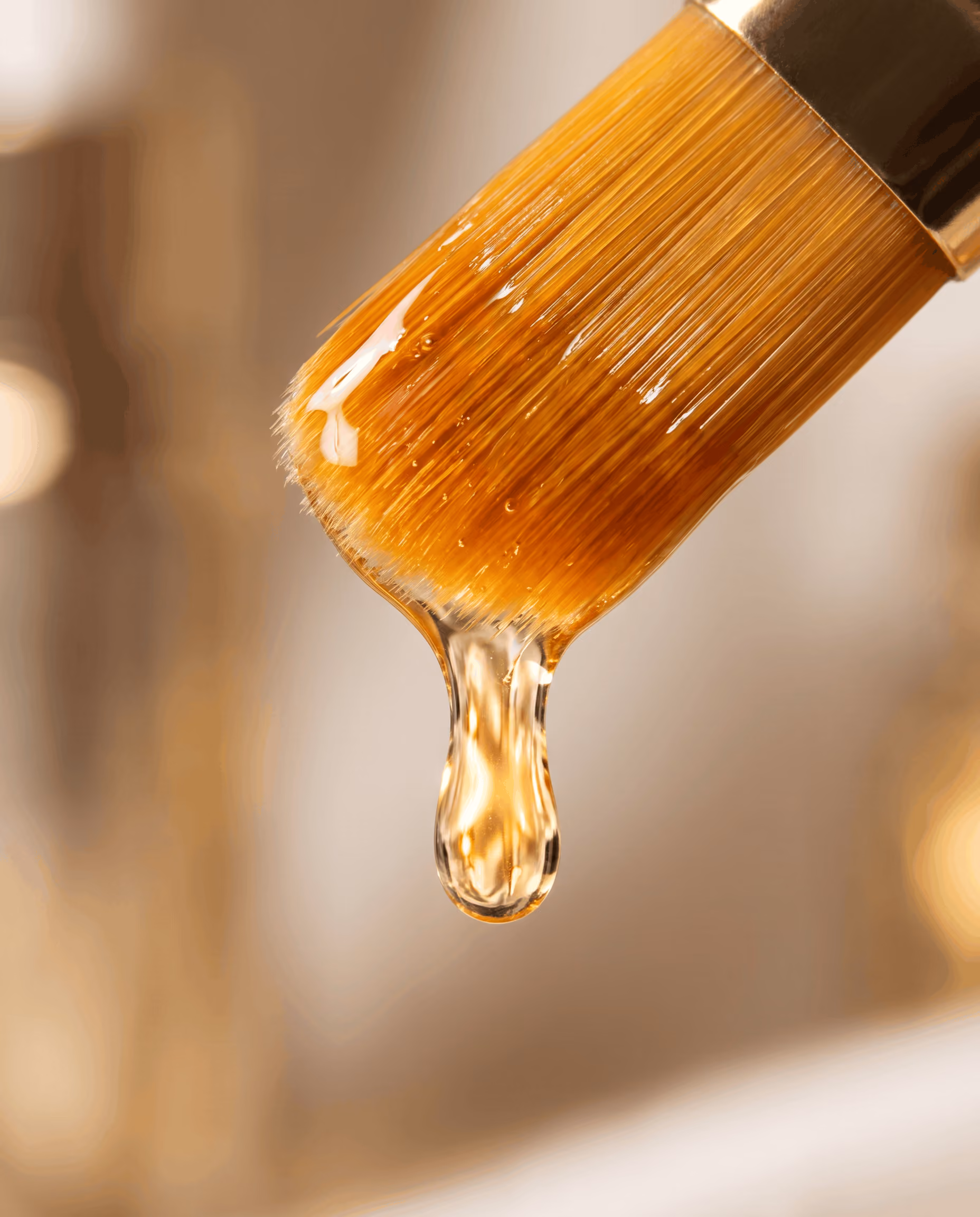 Close-up of a brush with golden-brown bristles dripping clear liquid, with a soft blurred background.