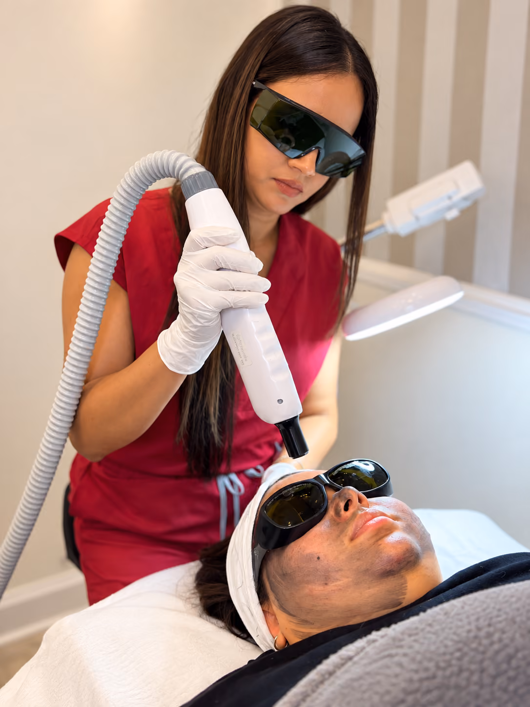 A woman in a red uniform wearing protective glasses performs a precision laser facial treatment on a client's face, who is lying down with protective goggles and a headband.