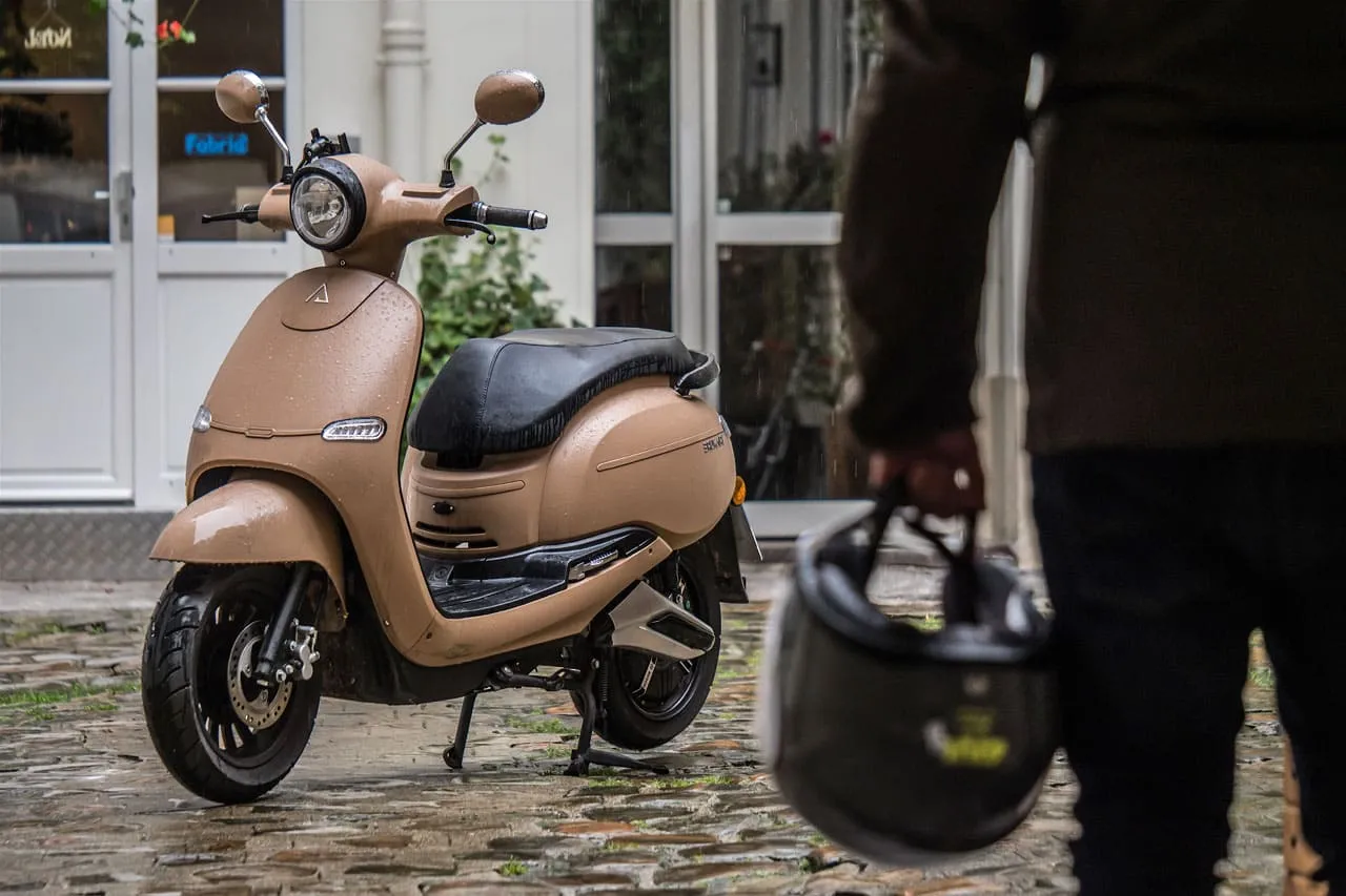 Scooter beige Brumaire à l'arrêt avec un homme à côté tenant un casque dans sa main