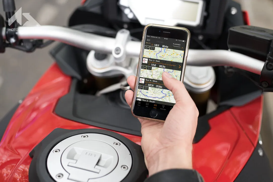 Une main de motard devant un guidon de moto qui tient un Smartphone où est ouvert l'application Rever