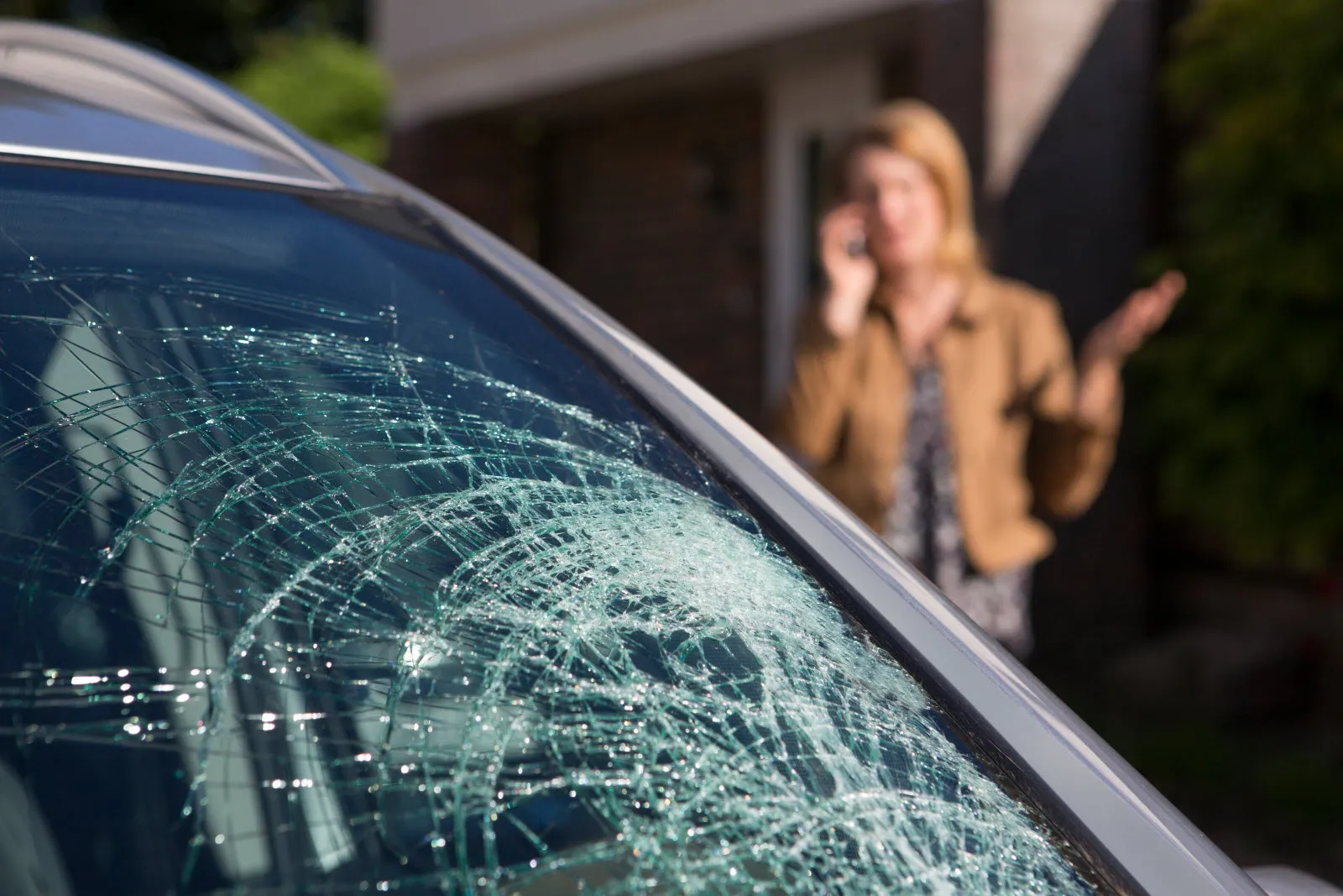 Un pare-brise de voiture cassé, avec en arrière plan une dame qui téléphone.