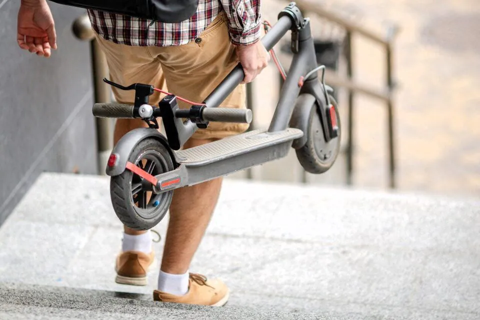 Un homme qui porte sa trottinette électrique pliable dans les mains en descendant un escalier.