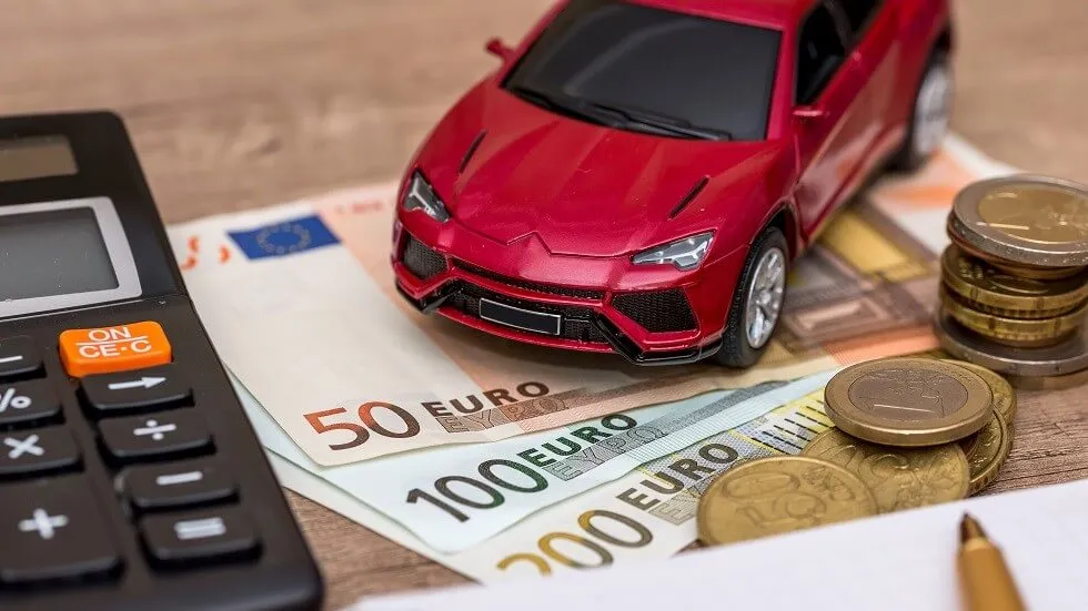 Une maquette de voiture rouge posé sur quelques liasses d'argent et des piles de monnaies, placées à côté d'une calculatrice.
