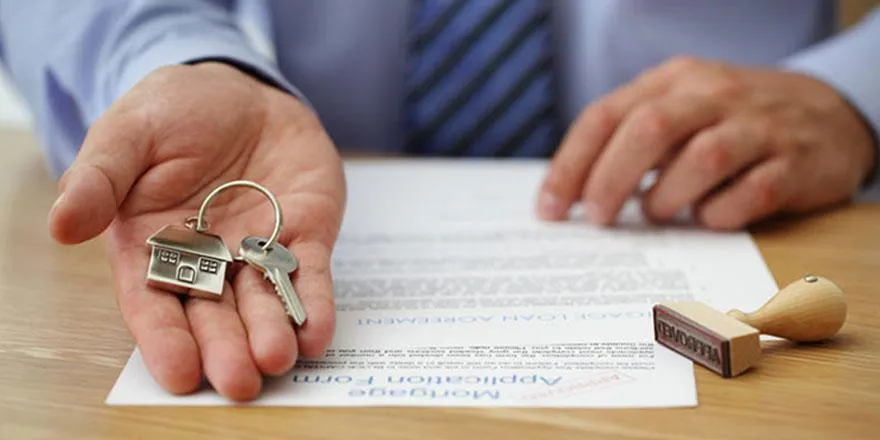Une main qui tient une clé avec un porte-clé en forme de maison. des documents et un tampon posé sur la table.