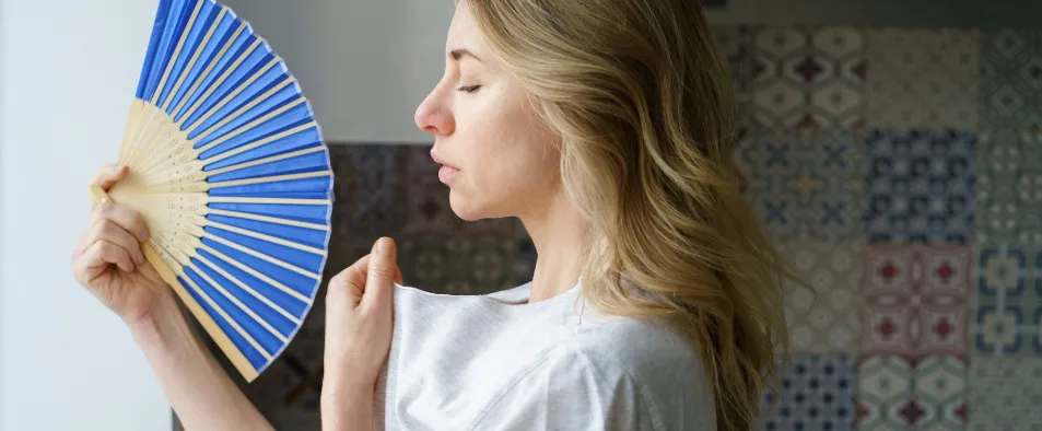 Une femme devant une fenêtre entrain de faire du vent avec son éventail sur son visage 