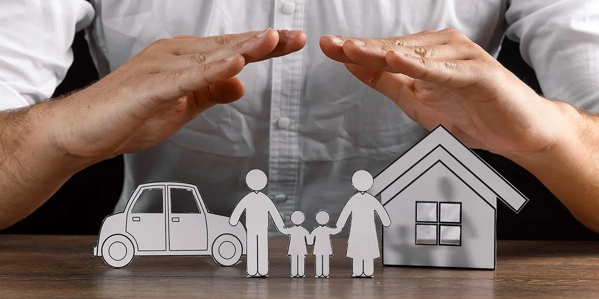 Des maquettes d'une voiture et d'une maison en papier avec une famille de deux adultes et deux enfants posées sur une table protégées par deux mains d'une personne qui forme un toit. 