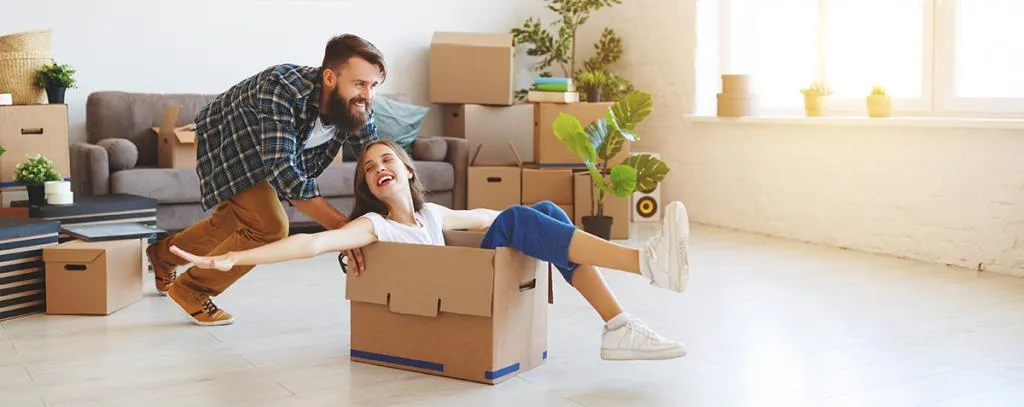 Un homme qui pousse une femme dans un carton de déménagement avec des cartons autour et un canapé à l'arrière