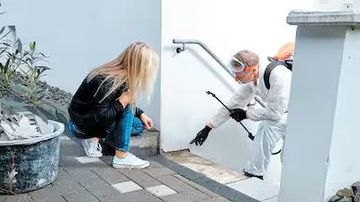 Une femme dans les escaliers qui discute avec un désinsectiseur professionnel dans une tenue blanche de protection.