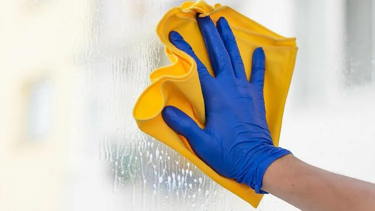 Une main qui porte un gant bleu et qui nettoie une vitre avec un chiffon jaune.