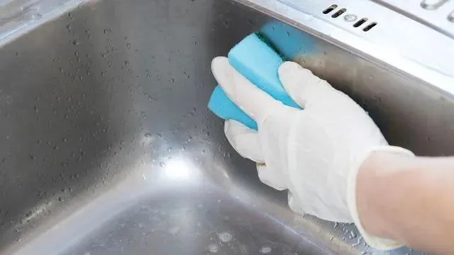 Une main qui porte un gant blanc et qui tient une éponge pour nettoyer un évier en inox.