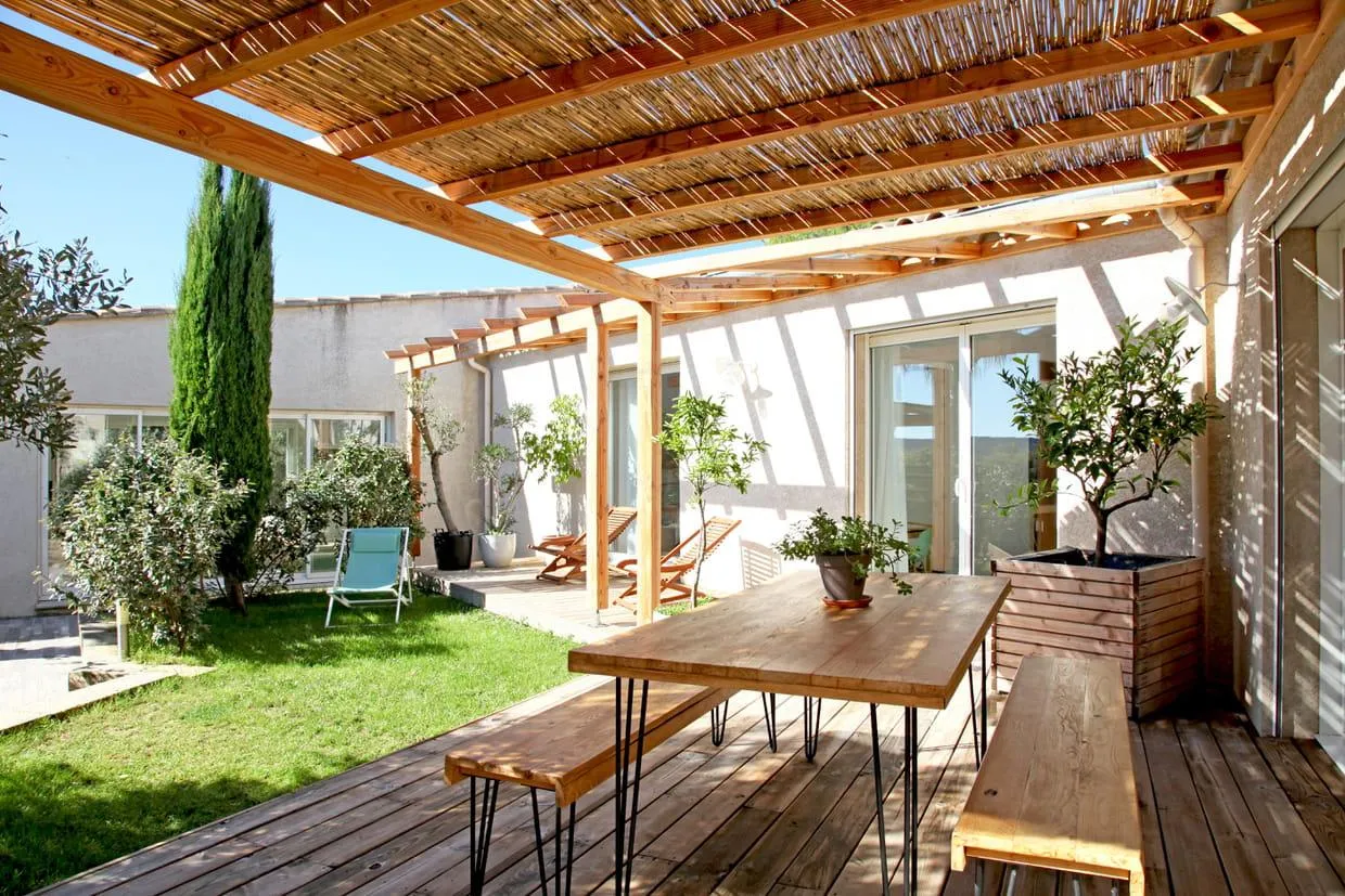 Une terrasse où se trouve une salle à manger d'extérieur avec une table et des chaises couvertes par des cannisses posées sur une pergola en bois.