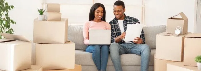Un couple installé sur un canapé entouré de plusieurs cartons. La femme est devant un ordinateur et l'homme tient des documents dans ses mains.