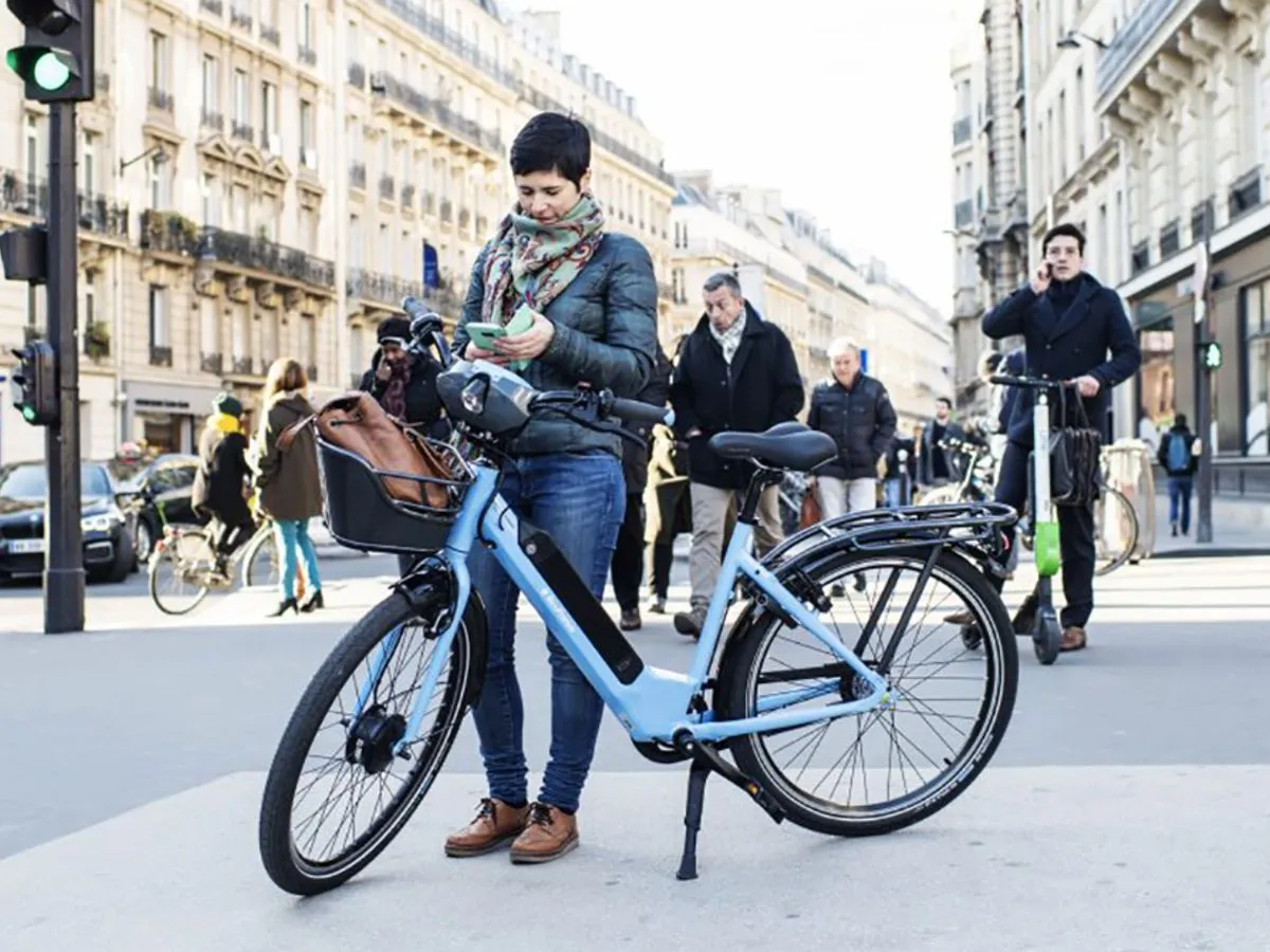 Une femme regardant son téléphone qui se tient à côté d'un vélo Véligo en stationnement en pleine rue