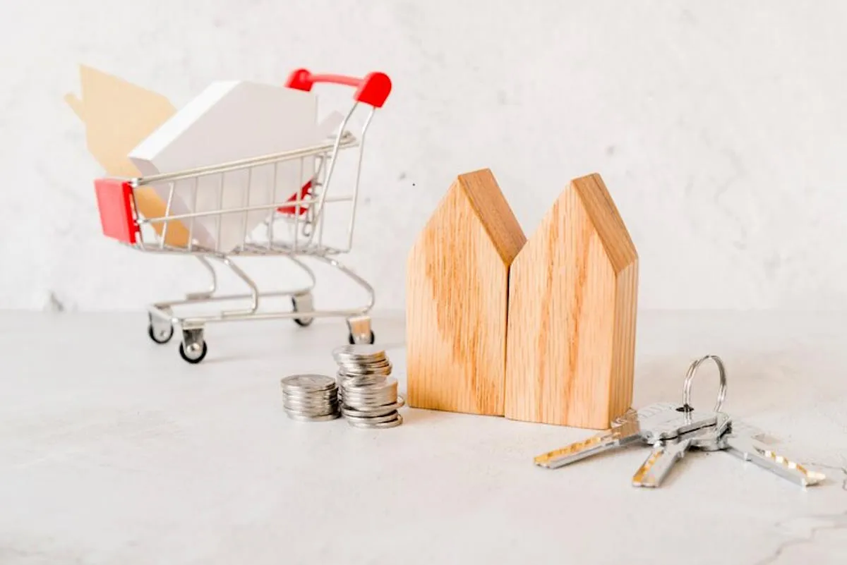 Une maquette de maison en bois posé à côté d’une pile de monnaies et de clés ainsi que d’un mini caddie