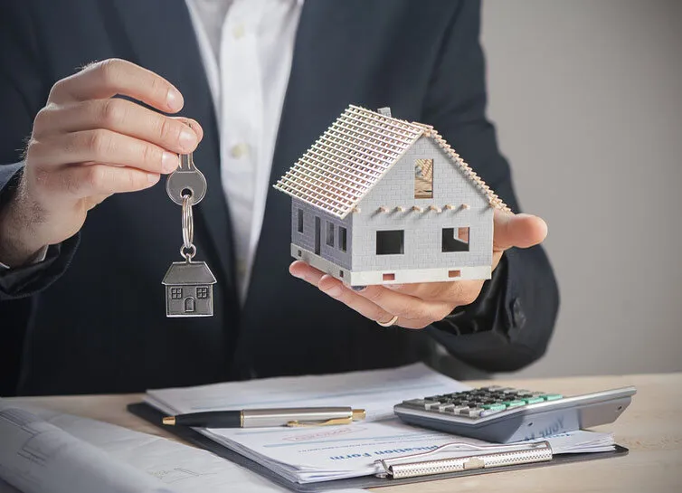 Un homme qui tient dans une main une clé et dans une autre une maquette de maison. Sur la table, il y a une calculatrice et des documents ainsi qu'un stylo