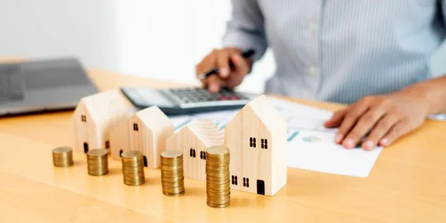 Une maquette de maison en bois avec des piles d'argent devant un homme qui utilise une machine à calculer sur un bureau