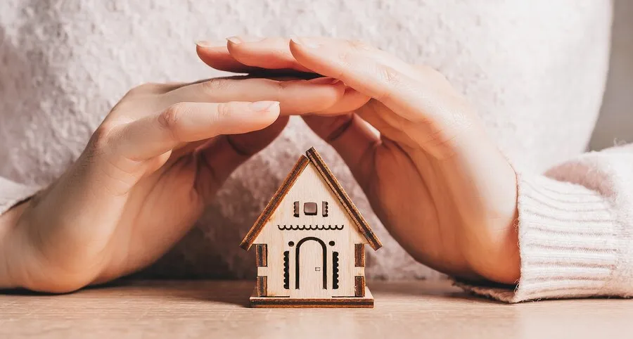 Une femme qui pose ses mains en forme de toit pour protéger une maquette de maison en bois posée sur une table