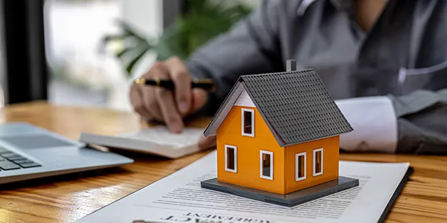 Une personne qui s'assoie devant une table et devant un ordinateur, une machine à calculer, une maquette de maison et un contrat d'assurance. 