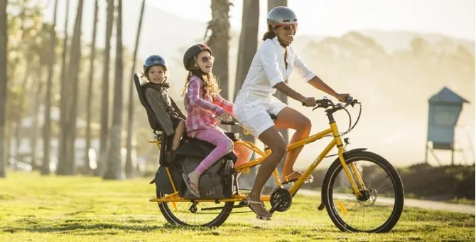 Une femme qui conduit ses enfants à bord d’un vélo longtail jaune et noir.