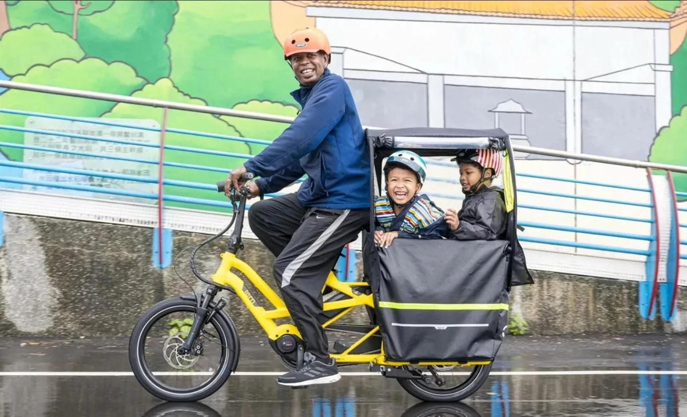 Un homme qui conduit ses deux garçons à bord d'un vélo longtail équipé d'une protection contre la pluie.