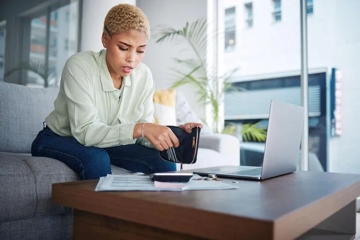 Une femme qui cherche quelque chose dans son portefeuille devant un ordinateur et des documents posés sur une table basse en bois