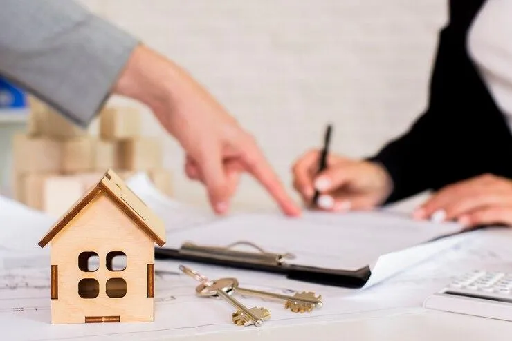 Une personne qui signe un document montré du doigts par un homme en arrière plant d'une maquette de maison et de clés posés sur une table