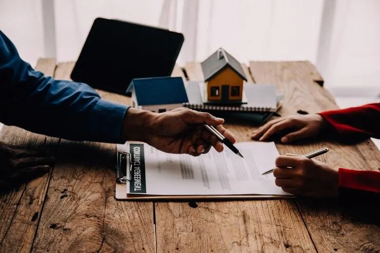 Une personne qui montre un contrat posé sur une table où il y a une maquette de maison et une tablette