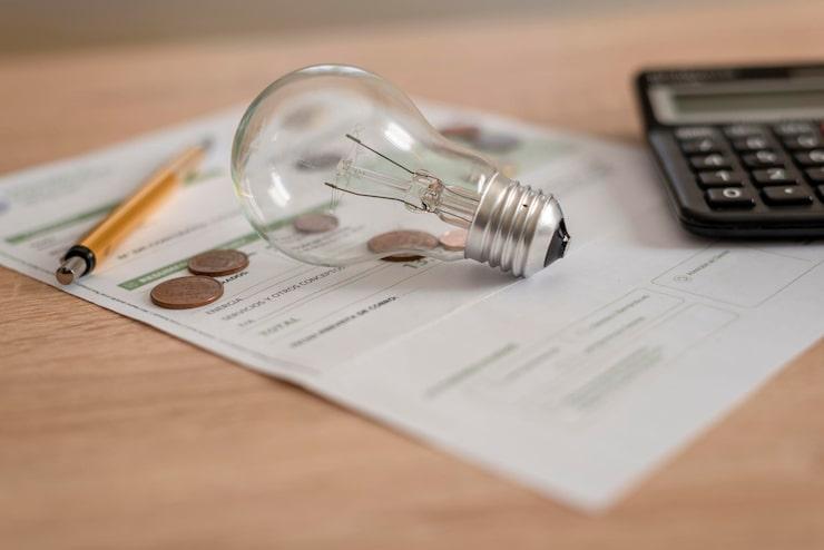 Une ampoule posée sur un document blanc à côté d'une calculatrice, des pièces de monnaies et un stylo