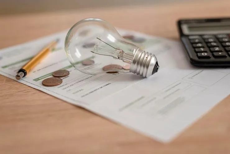 Une ampoule posée sur un document blanc à côté d'une calculatrice, des pièces de monnaies et un stylo