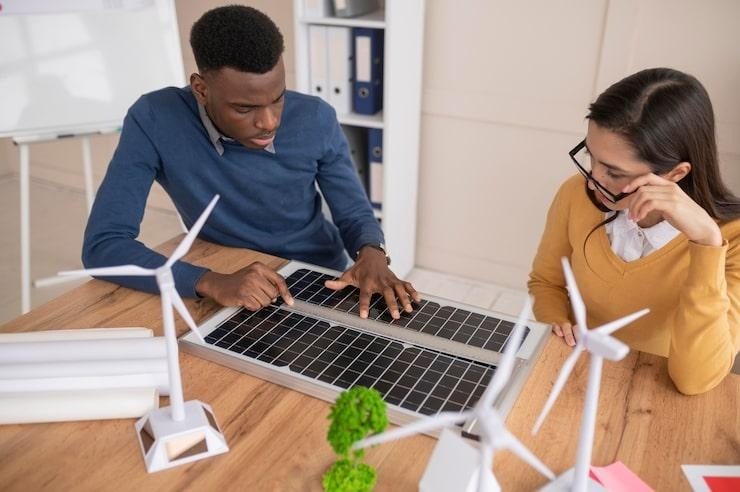 Un homme et une femme qui regarde un panneau solaire pos sur une table où sont aussi posés des maquettes d'éoliennes