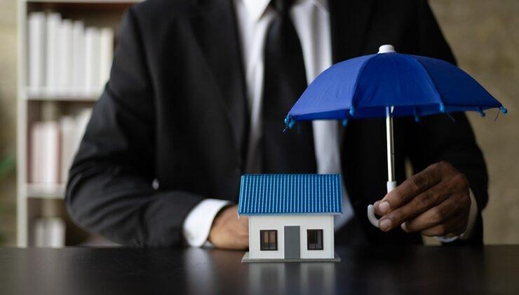 Un homme qui tient un parapluie bleu pour protéger une maquette de maison posée sur une table