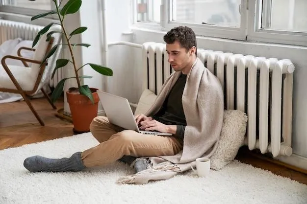 Un homme installé sur le sol contre un radiateur et qui porte une couverture en travaillant sur son ordinateur