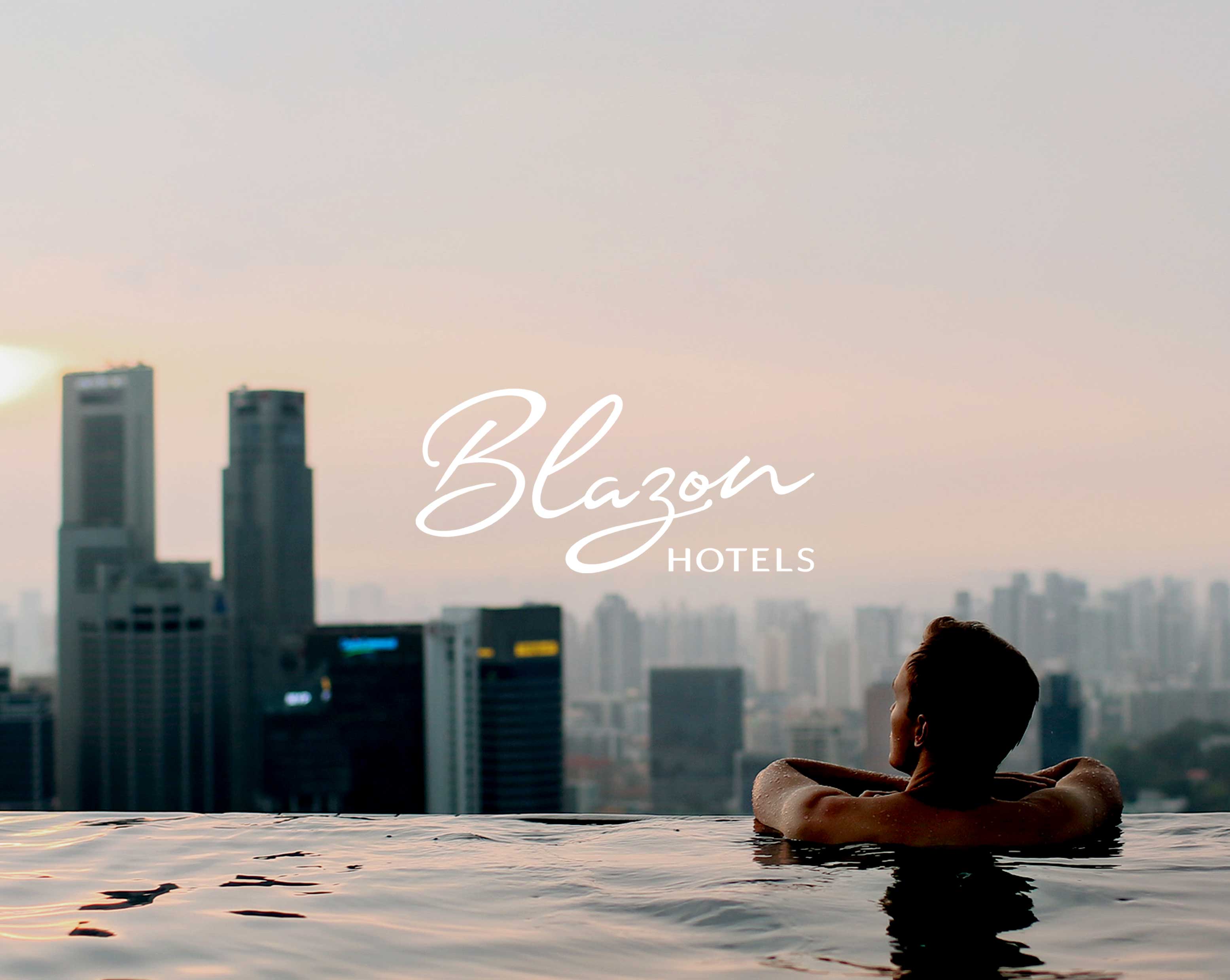 Person relaxing in an infinity pool overlooking a city skyline at sunset with Blazon Hotels logo overlay.