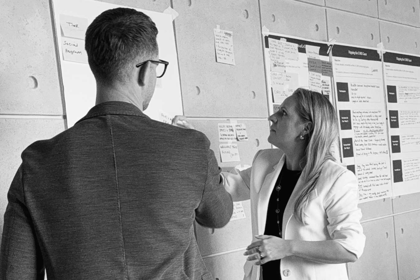 Two professionals reviewing notes and sticky notes on a wall during a planning session.