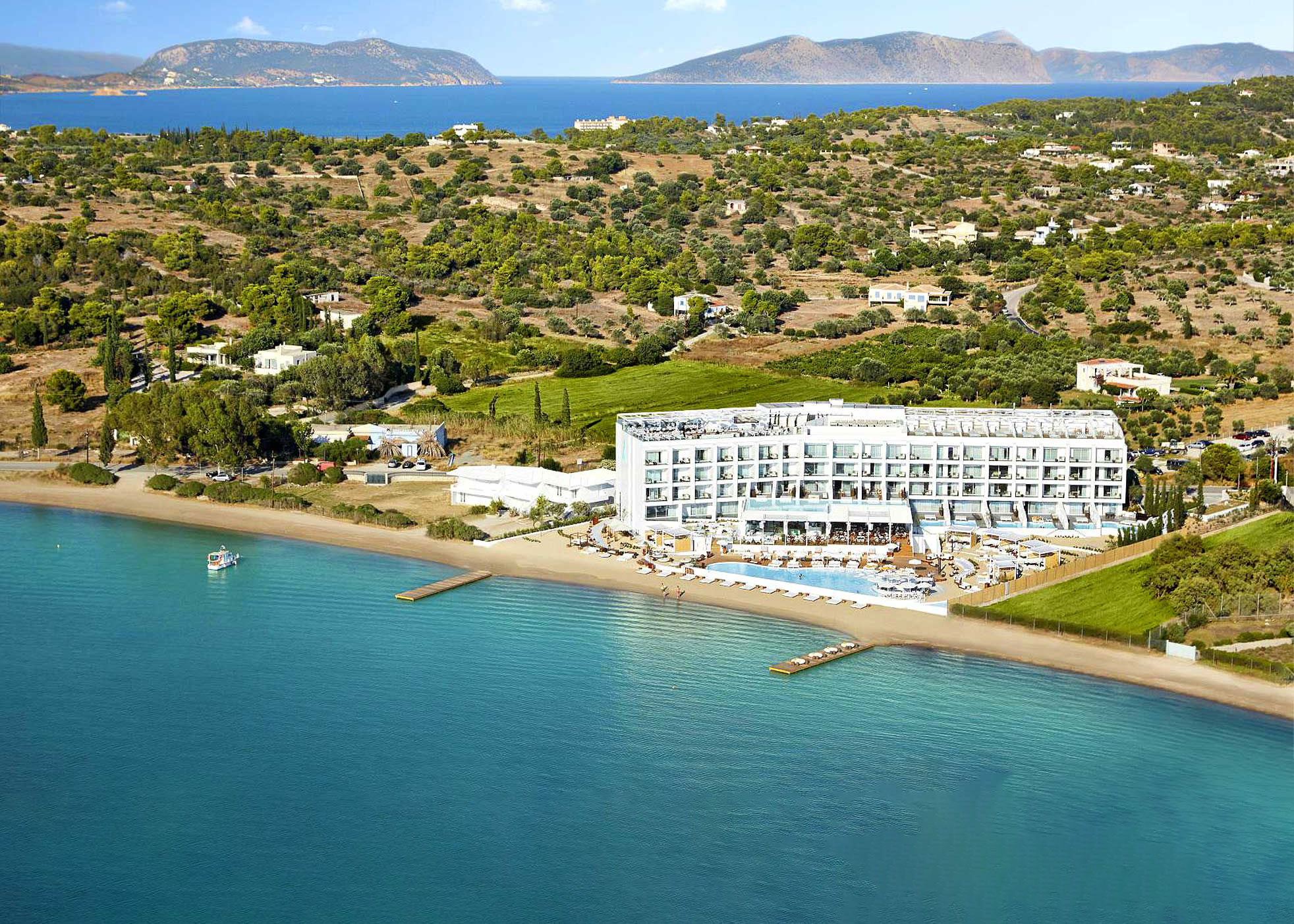 Beachfront hotel with pool and lounge chairs by clear blue sea, surrounded by green hills and mountains.