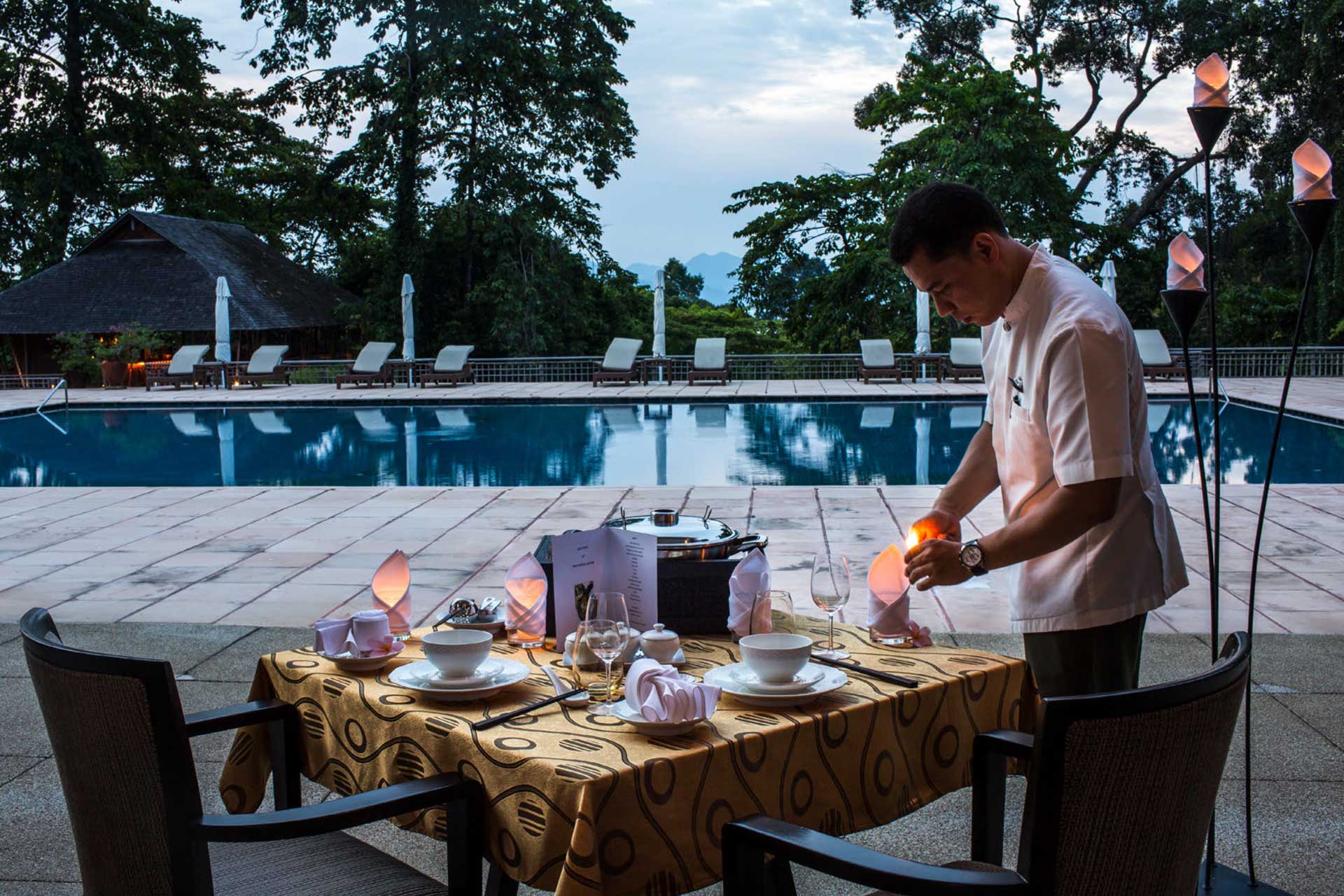 Brandteliers Singapore is a Luxury Hospitality Branding agency and a photograph of a waiter lighting a candle on a set dining table beside a calm outdoor pool with lounge chairs and trees in the background.