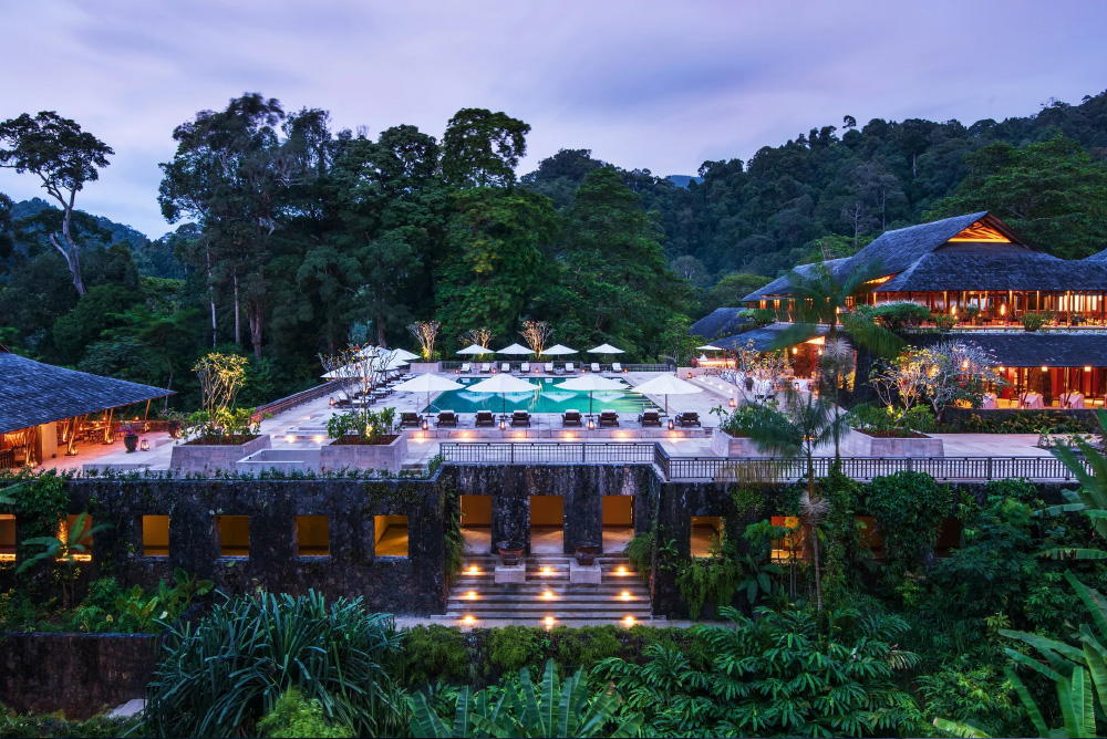 Brandteliers Singapore is a Luxury Hospitality Branding agency and a photograph of a Luxury resort in Malaysia at dusk with illuminated swimming pool surrounded by lounge chairs, umbrellas, and tropical forest.