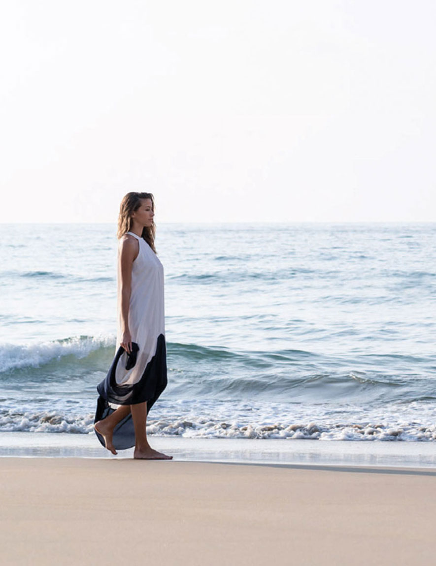Woman in a flowing white and black dress walking barefoot on a sandy beach near gentle ocean waves.