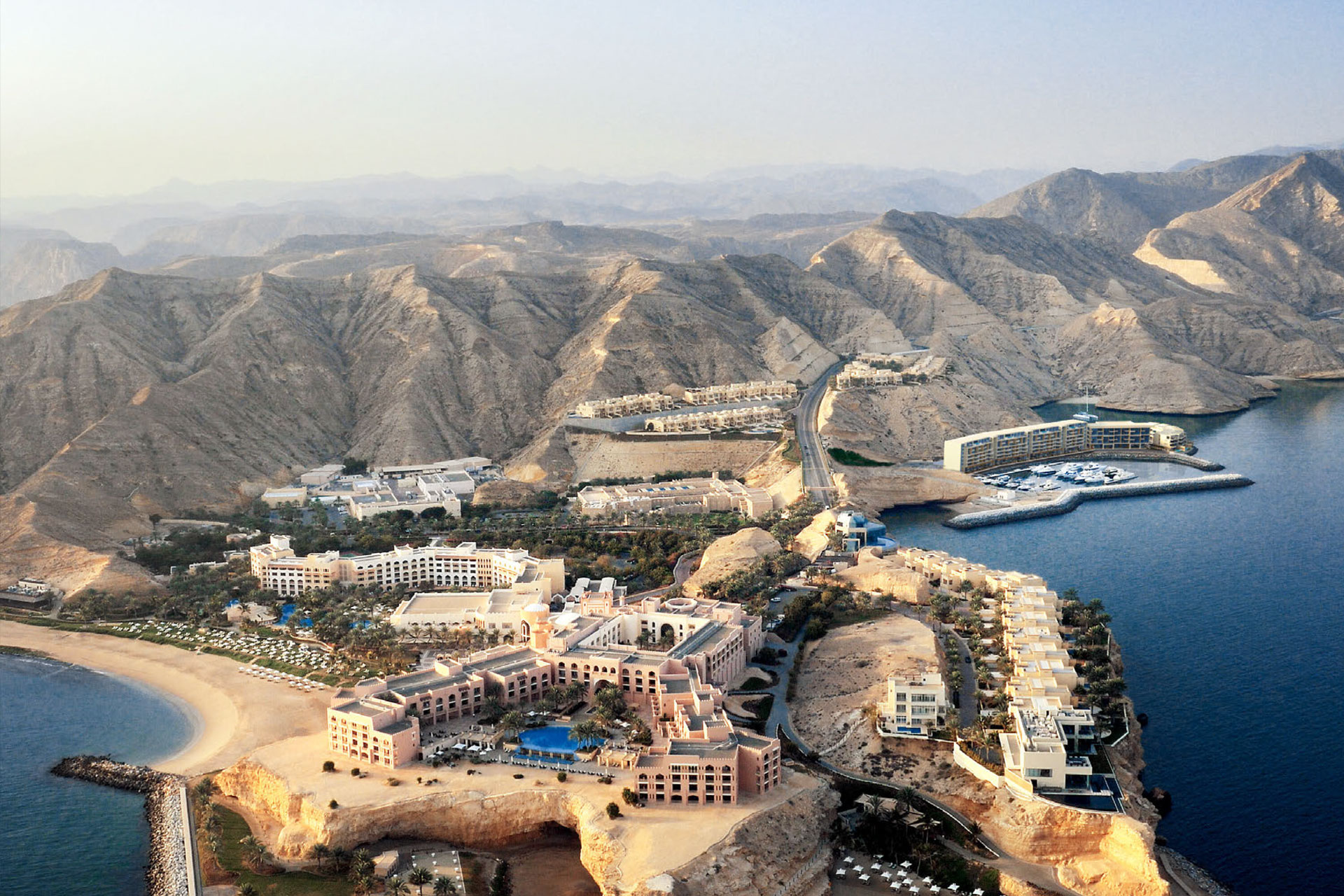 Aerial view of a coastal resort complex with multiple buildings, swimming pools, and a marina surrounded by rugged mountains and calm sea.