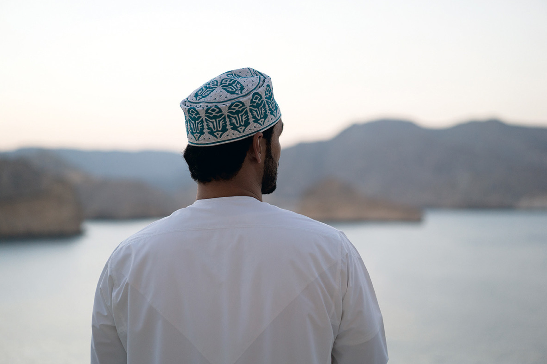 Man in traditional white clothing and embroidered cap looking at mountains across a body of water.